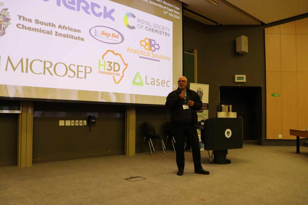 A person stands on a stage presenting to an audience. The large screen behind them displays logos of sponsors and collaborators.