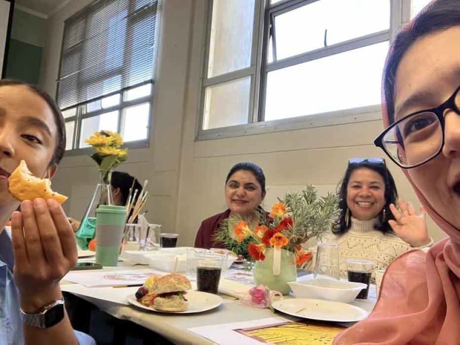 A group of people sitting around a table with food, drinks, and a vase of yellow flowers. The setting is an indoor room with windows and blinds in the background.