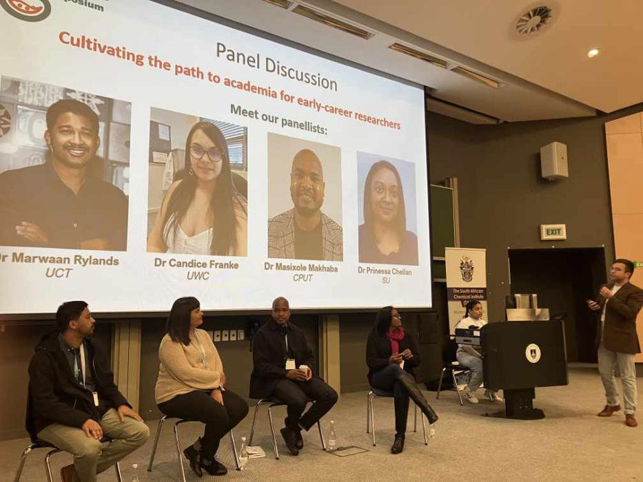 Five panelists are seated at the front, with their faces obscured for privacy. Above them, a projected screen displays the title of the event and introduces four of the panelists with their names, titles, and affiliations. A person is standing on the right side addressing the audience with a microphone in hand.