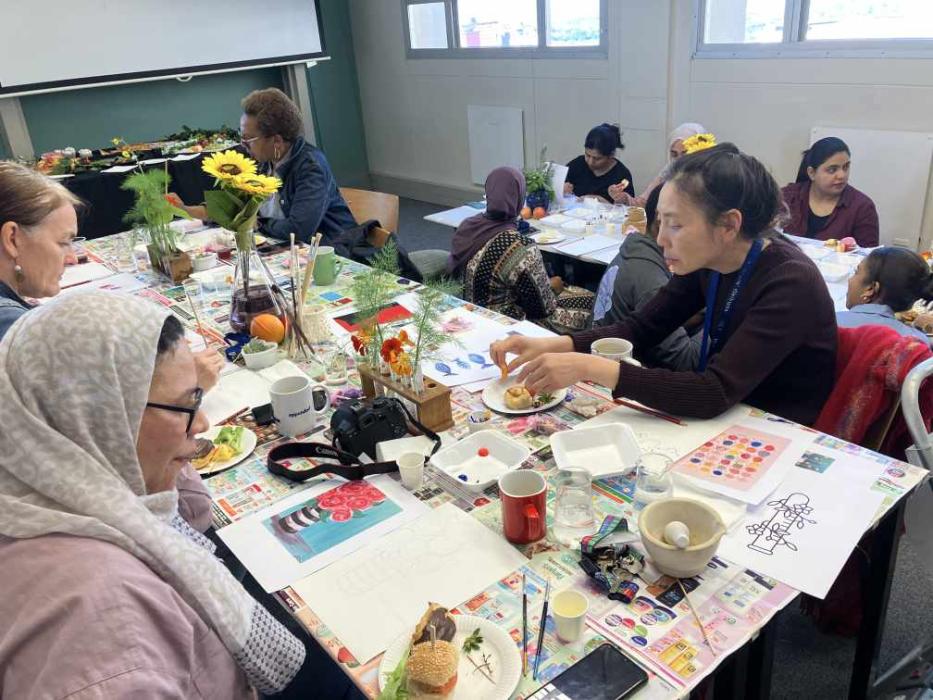 A group of individuals engaged in an art class. The tables are covered with various art supplies, including paint palettes, brushes, and watercolor papers. Some participants are painting, while others are handling different materials or having a snack. The room is bright with natural light coming in from the windows.