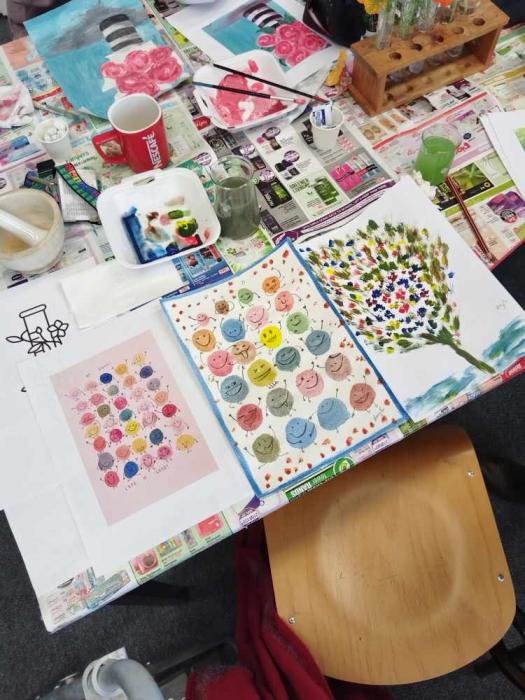 A group of people engaged in a painting activity around a table, with various art supplies like paintbrushes, paints, and palettes visible.