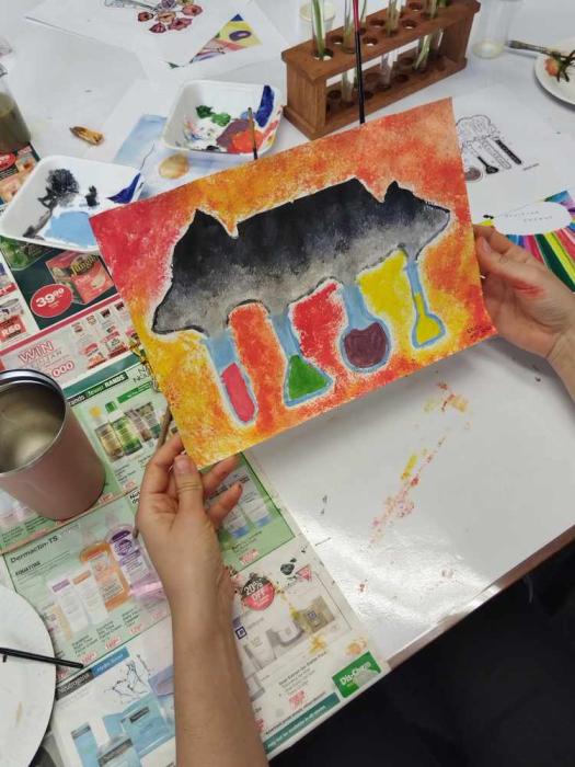 A pair of hands holding a colorful painting with an abstract design featuring a black silhouette resembling an animal, possibly a cow, with vibrant patterns and drips below. The background includes a table covered with newspapers and art supplies, suggesting an art class or creative workshop.
