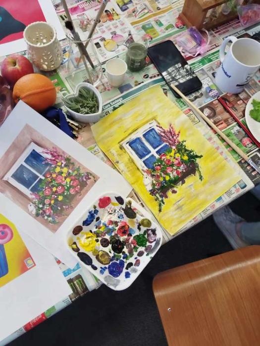 A cluttered table with a watercolor painting of a window with flowers, an open book, paintbrushes, a palette with multiple colors of paint, cups, a smartphone, fruit, and other miscellaneous items, suggesting a creative workspace.