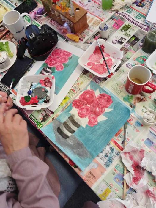 A painting in progress on a canvas, depicting pink roses in a striped vase, surrounded by painting supplies including brushes, a palette with paint, tissues, and two mugs, suggesting an art workshop or personal creative space.