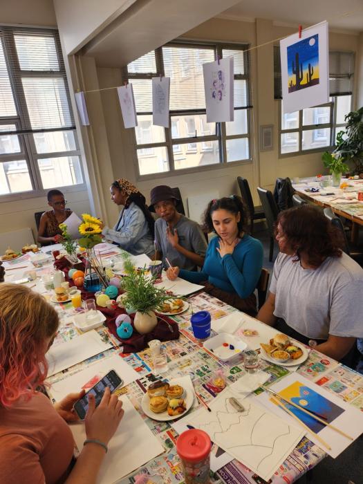 A group of individuals sitting around a table covered with newspapers, art supplies, and food items like fruits and sandwiches. A vase with sunflowers is in the center. Some people are crafting, while others are eating or conversing. The room has posters on the walls and large windows letting in natural light.