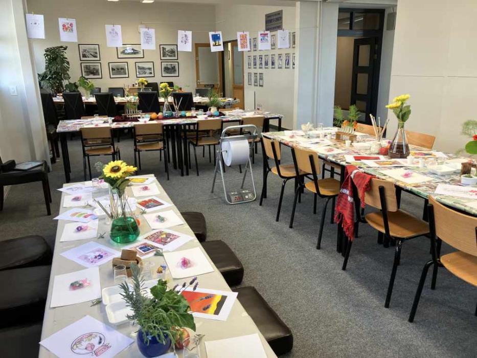 An art workshop with tables arranged in a U-shape, covered with paintbrushes, palettes, and watercolor papers. The room is colorful, with paintings on the walls and easels. A green vase with yellow flowers is in the center.