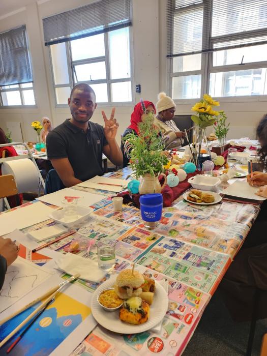 A classroom setting with students sitting around a table covered with a colorful tablecloth, various craft materials, and food items. A plate with food is in the foreground, and students are engaged in activities. Faces are not visible or are obscured for privacy.