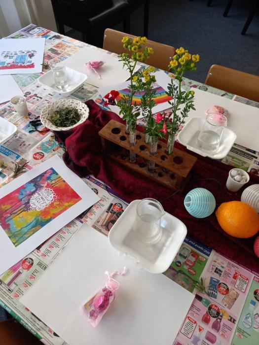 A table set up for a craft or educational activity with a wooden block holding colorful flowers, teacups, a teapot, greenery, paint supplies, printed pictures, oranges, and a blue paper lantern on a tablecloth with text and images.