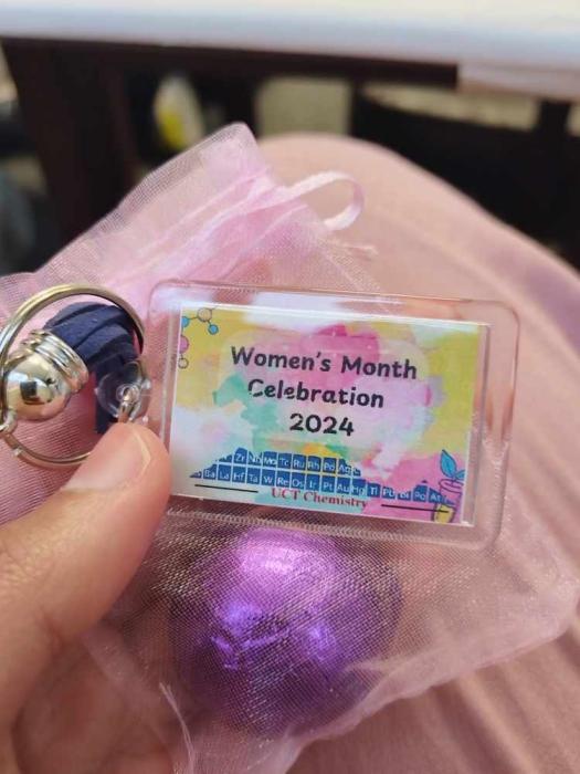 A hand holding a small, rectangular badge with a keychain. The badge reads ‘Women’s Month Celebration 2024’ with colorful abstract designs and a chemistry-related logo. The badge is on a pink fabric surface.
