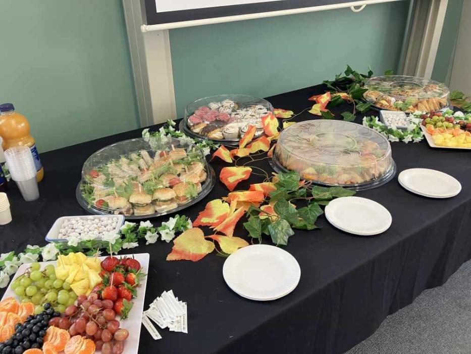 A buffet table set up for an event with platters of sandwiches, wraps, fruits, and vegetables. Decorative foliage with autumn leaves accents the presentation. A chalkboard and a bottle of orange juice are in the background.