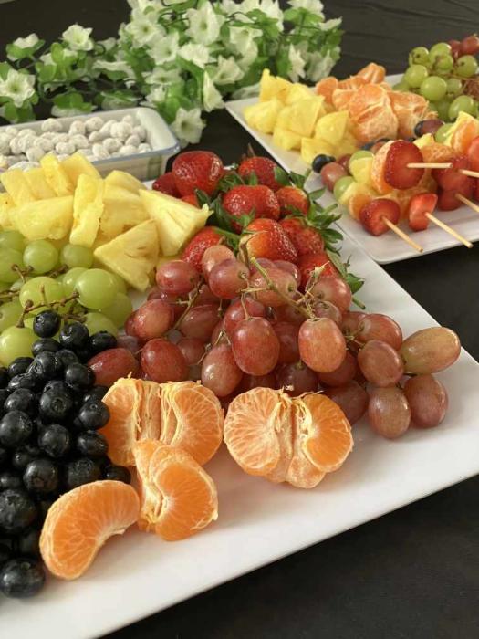 A white platter filled with a variety of fresh fruits, including red and green grapes, pineapple slices, strawberries, blackberries, and mandarin orange segments. In the background, there are fruit skewers and a bowl of nuts or small snacks on a dark surface with white flowers in the upper left corner.