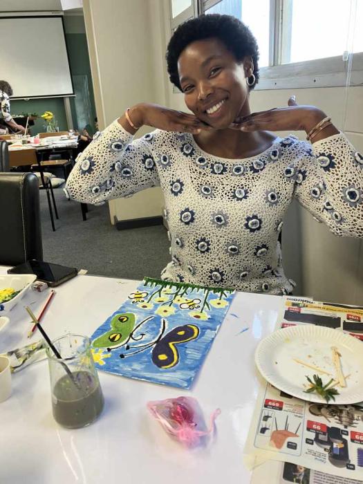 An individual is sitting at a table with a painting of a blue butterfly with yellow and green details on a background of green grass and blue sky, with yellow flowers at the bottom. The table has paintbrushes, a jar of water, and various colors of paint, suggesting an art class or workshop setting.