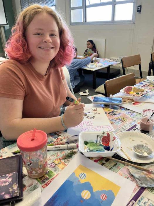 An individual is sitting at a table covered with newspapers, engaged in painting. Various art supplies, including paint palettes and brushes, are scattered around. In front of the individual is a colorful artwork featuring blue and yellow shapes on paper, suggesting an art class or workshop setting.