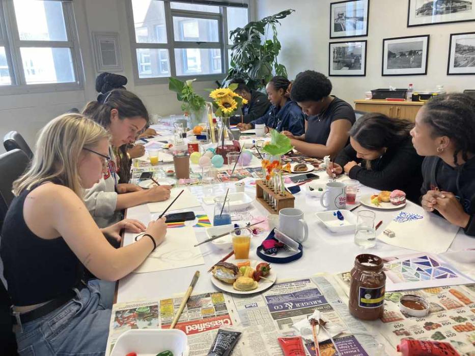 A group of individuals engaged in an art activity around a table covered with newspapers. The table has a vase of sunflowers, various painting supplies, personal items, beverages, and snacks, indicating a casual and social atmosphere.