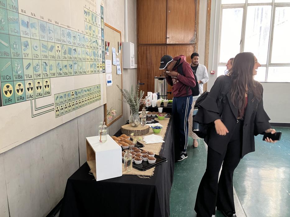 Classroom with students viewing a periodic table and a table with baked goods, likely during a science event.
