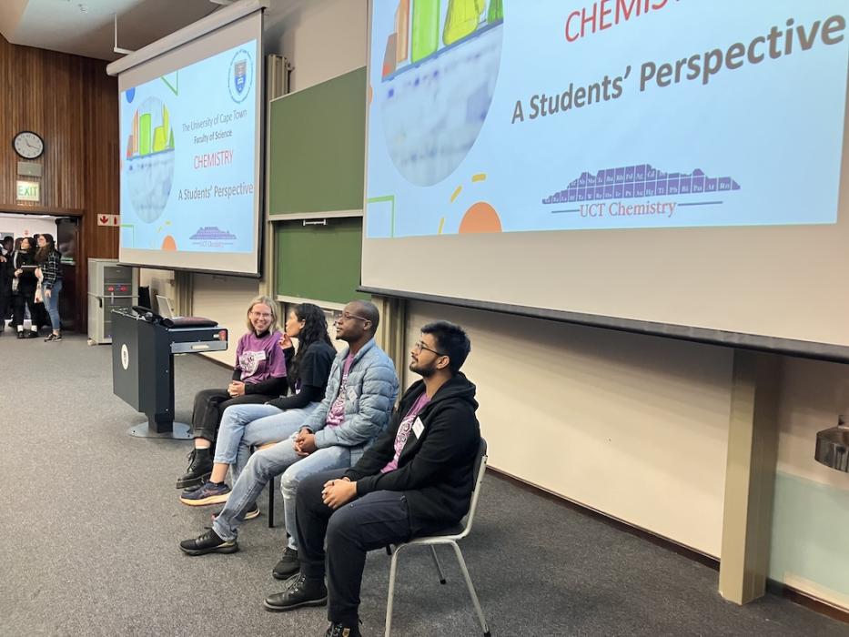 Four chemistry department postgraduate students sit on chairs in front of lecture theatre boards