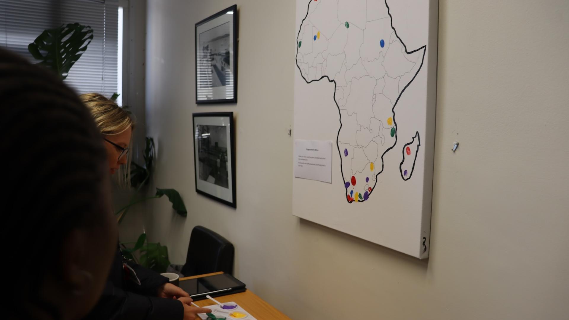 An outline of Afrika on a white canvas that is hung up on the wall with six colours paint on the table for students to show where in Afrika they come from.