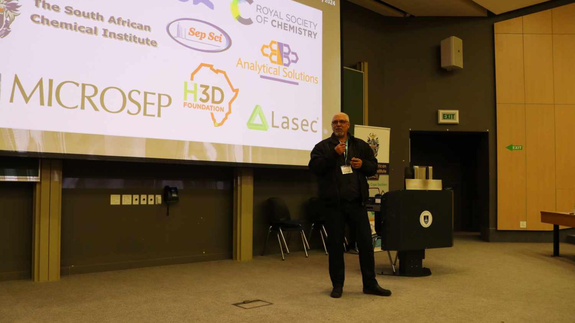 A person stands on a stage presenting to an audience. The large screen behind them displays logos of sponsors and collaborators.