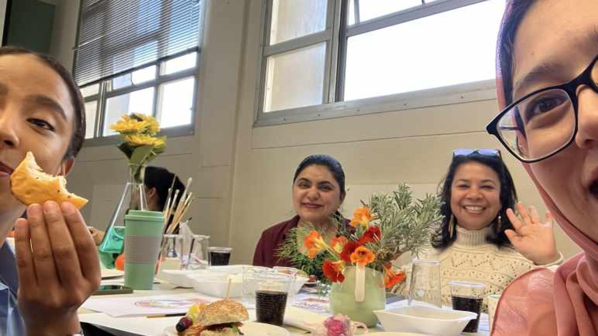 A group of people sitting around a table with food, drinks, and a vase of yellow flowers. The setting is an indoor room with windows and blinds in the background.