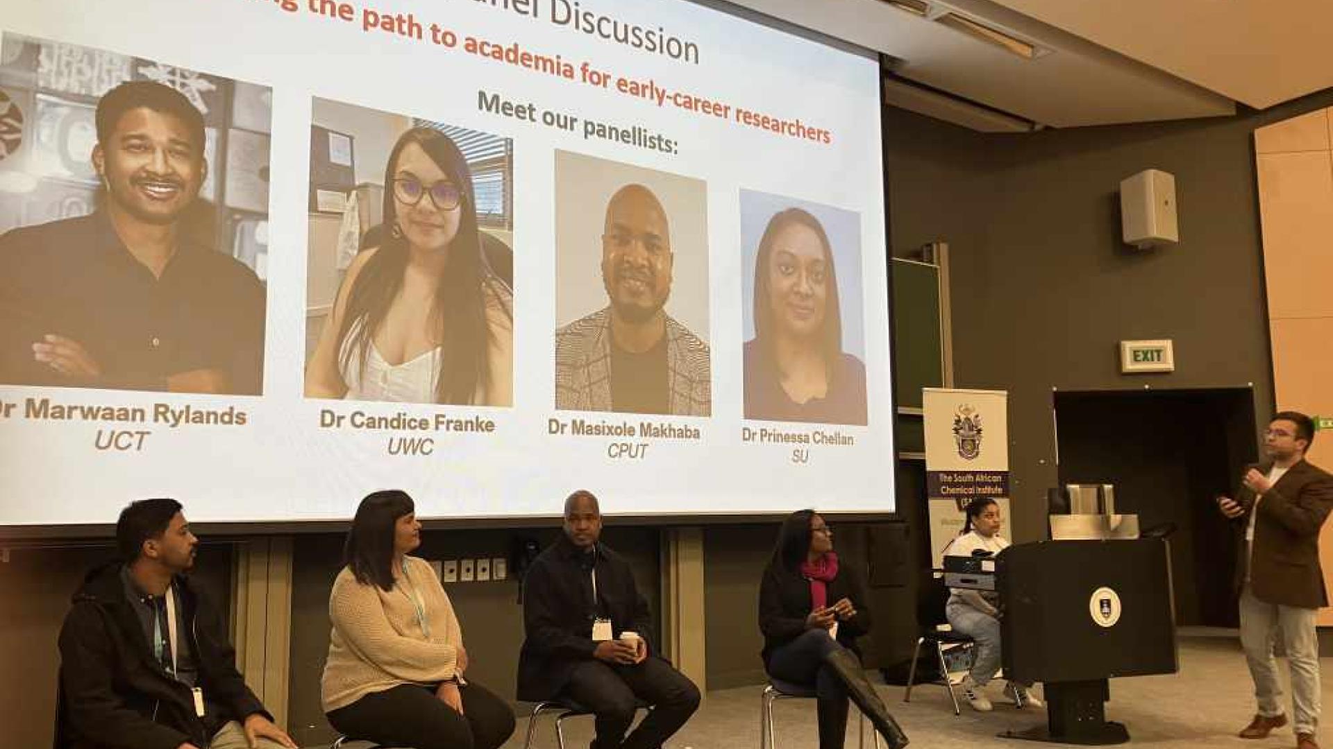 Five panelists are seated at the front, with their faces obscured for privacy. Above them, a projected screen displays the title of the event and introduces four of the panelists with their names, titles, and affiliations. A person is standing on the right side addressing the audience with a microphone in hand.