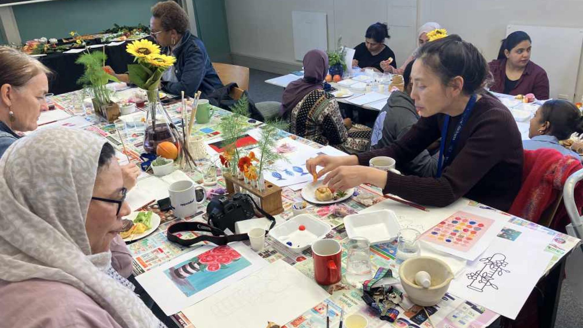A group of individuals engaged in an art class. The tables are covered with various art supplies, including paint palettes, brushes, and watercolor papers. Some participants are painting, while others are handling different materials or having a snack. The room is bright with natural light coming in from the windows.