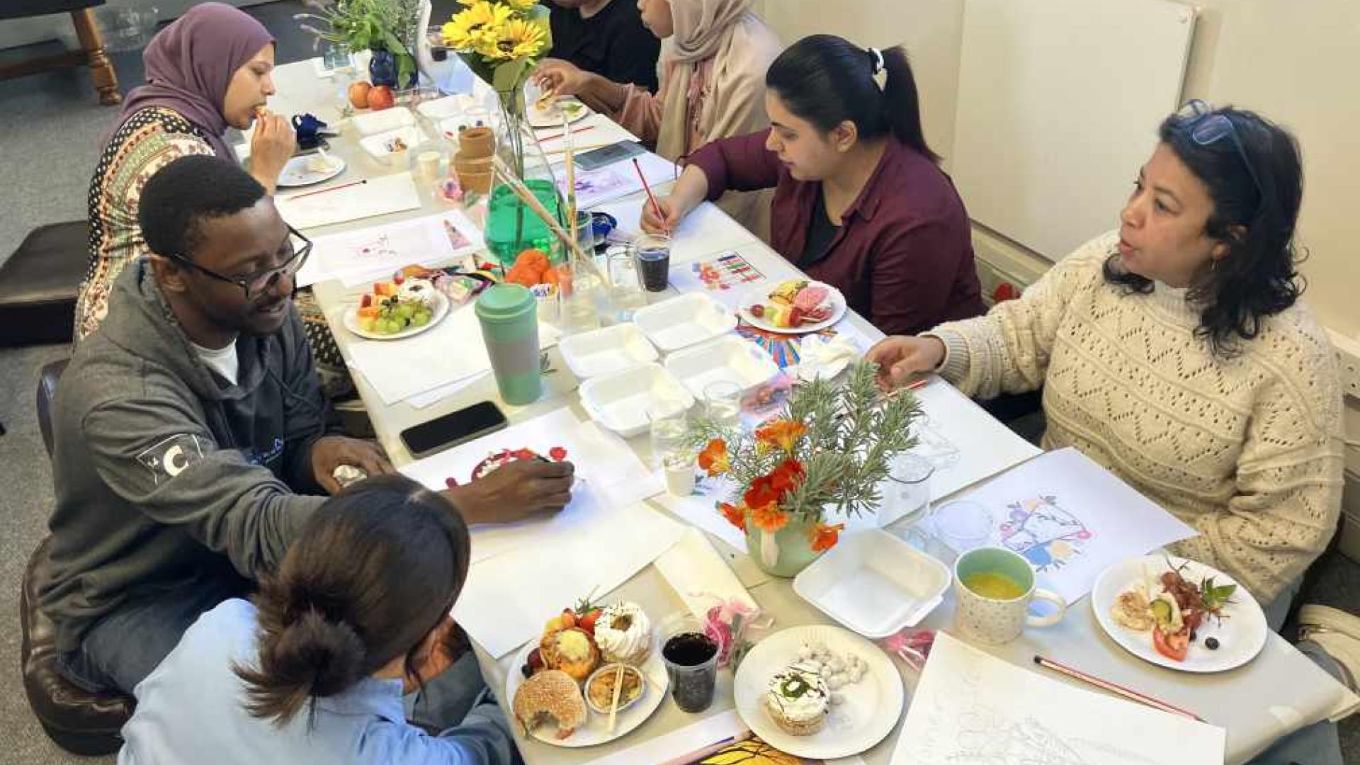 A group of people seated around a table with various dishes, beverages, and colorful flowers, in a casual dining setting.