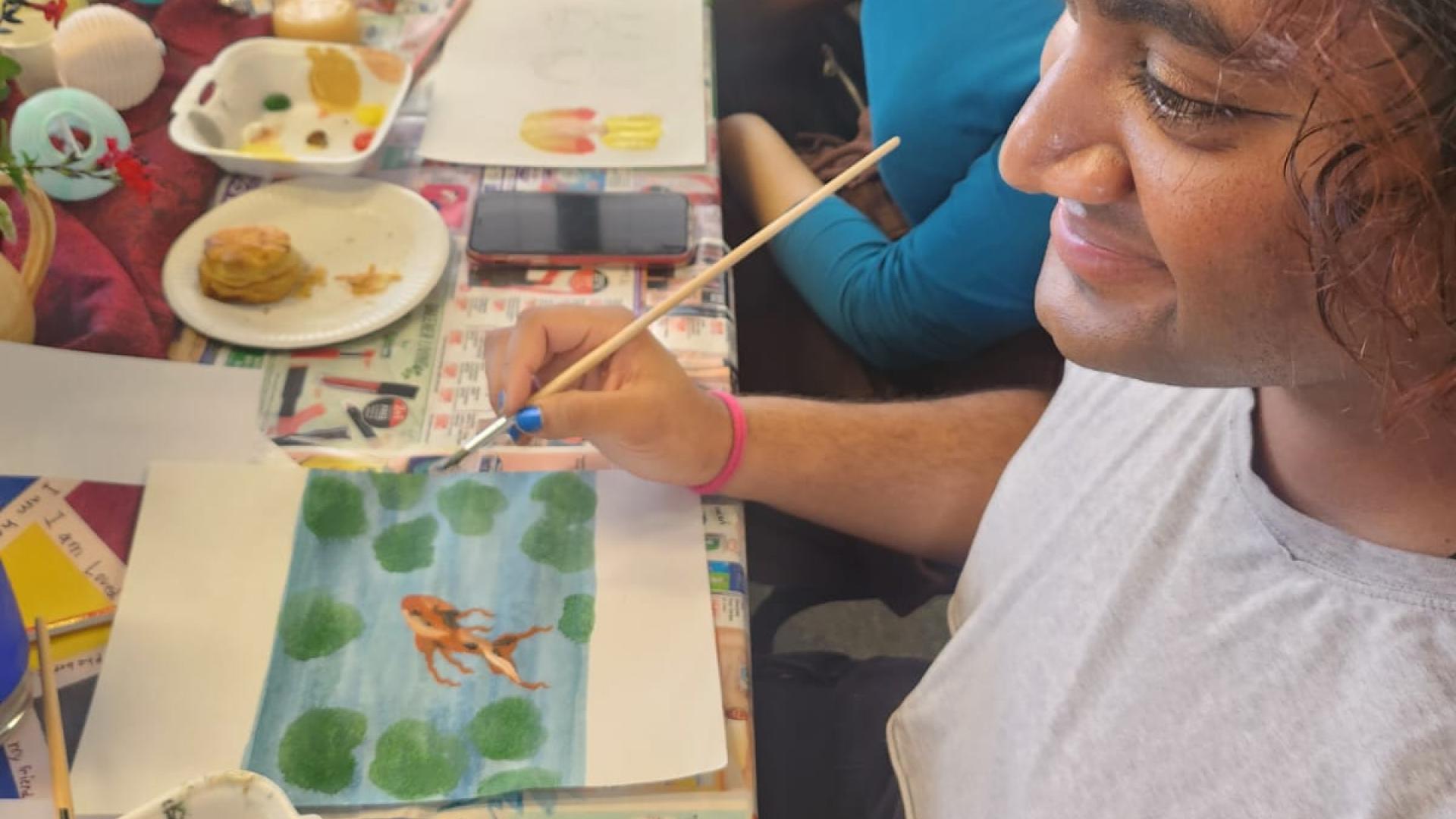 An artist’s workspace with various art supplies, including paint blobs on sheets of paper, a palette with different colors, brushes, a cup of water, and a wooden stool.
