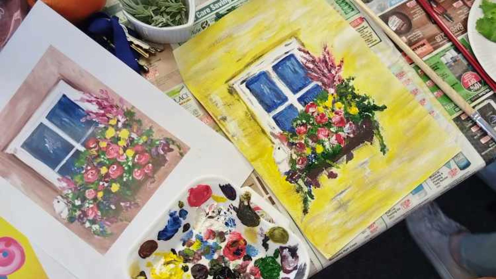 A cluttered table with a watercolor painting of a window with flowers, an open book, paintbrushes, a palette with multiple colors of paint, cups, a smartphone, fruit, and other miscellaneous items, suggesting a creative workspace.
