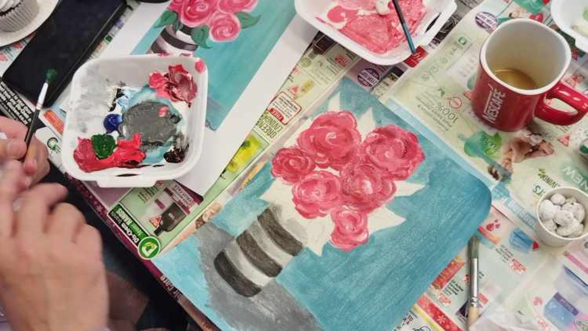 A painting in progress on a canvas, depicting pink roses in a striped vase, surrounded by painting supplies including brushes, a palette with paint, tissues, and two mugs, suggesting an art workshop or personal creative space.