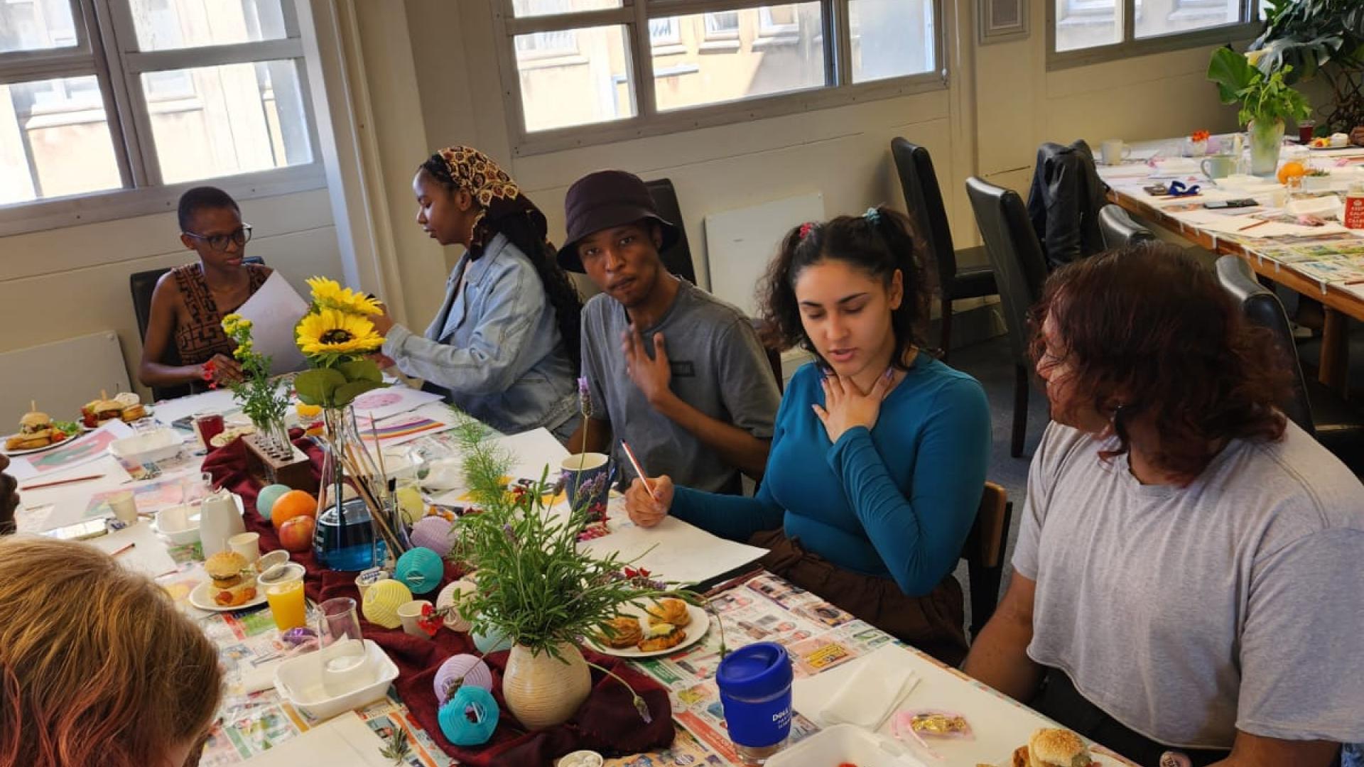 A group of individuals sitting around a table covered with newspapers, art supplies, and food items like fruits and sandwiches. A vase with sunflowers is in the center. Some people are crafting, while others are eating or conversing. The room has posters on the walls and large windows letting in natural light.