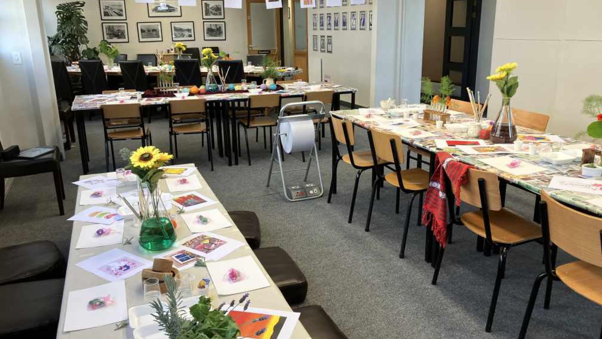 An art workshop with tables arranged in a U-shape, covered with paintbrushes, palettes, and watercolor papers. The room is colorful, with paintings on the walls and easels. A green vase with yellow flowers is in the center.