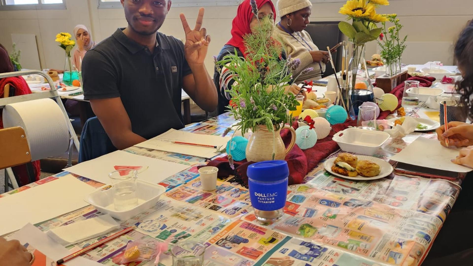 A classroom setting with students sitting around a table covered with a colorful tablecloth, various craft materials, and food items. A plate with food is in the foreground, and students are engaged in activities. Faces are not visible or are obscured for privacy.
