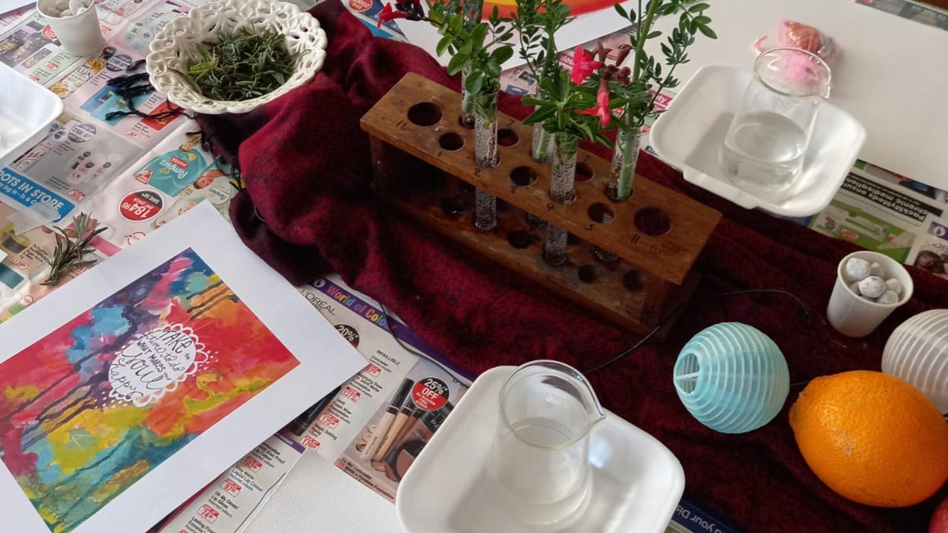 A table set up for a craft or educational activity with a wooden block holding colorful flowers, teacups, a teapot, greenery, paint supplies, printed pictures, oranges, and a blue paper lantern on a tablecloth with text and images.