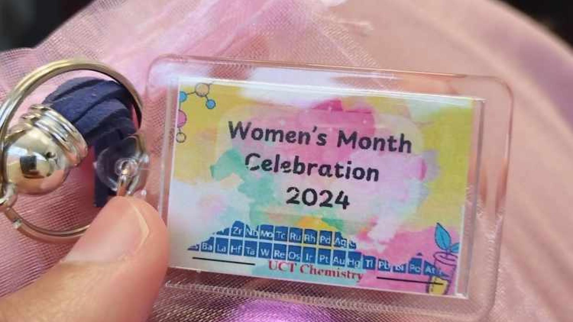 A hand holding a small, rectangular badge with a keychain. The badge reads ‘Women’s Month Celebration 2024’ with colorful abstract designs and a chemistry-related logo. The badge is on a pink fabric surface.