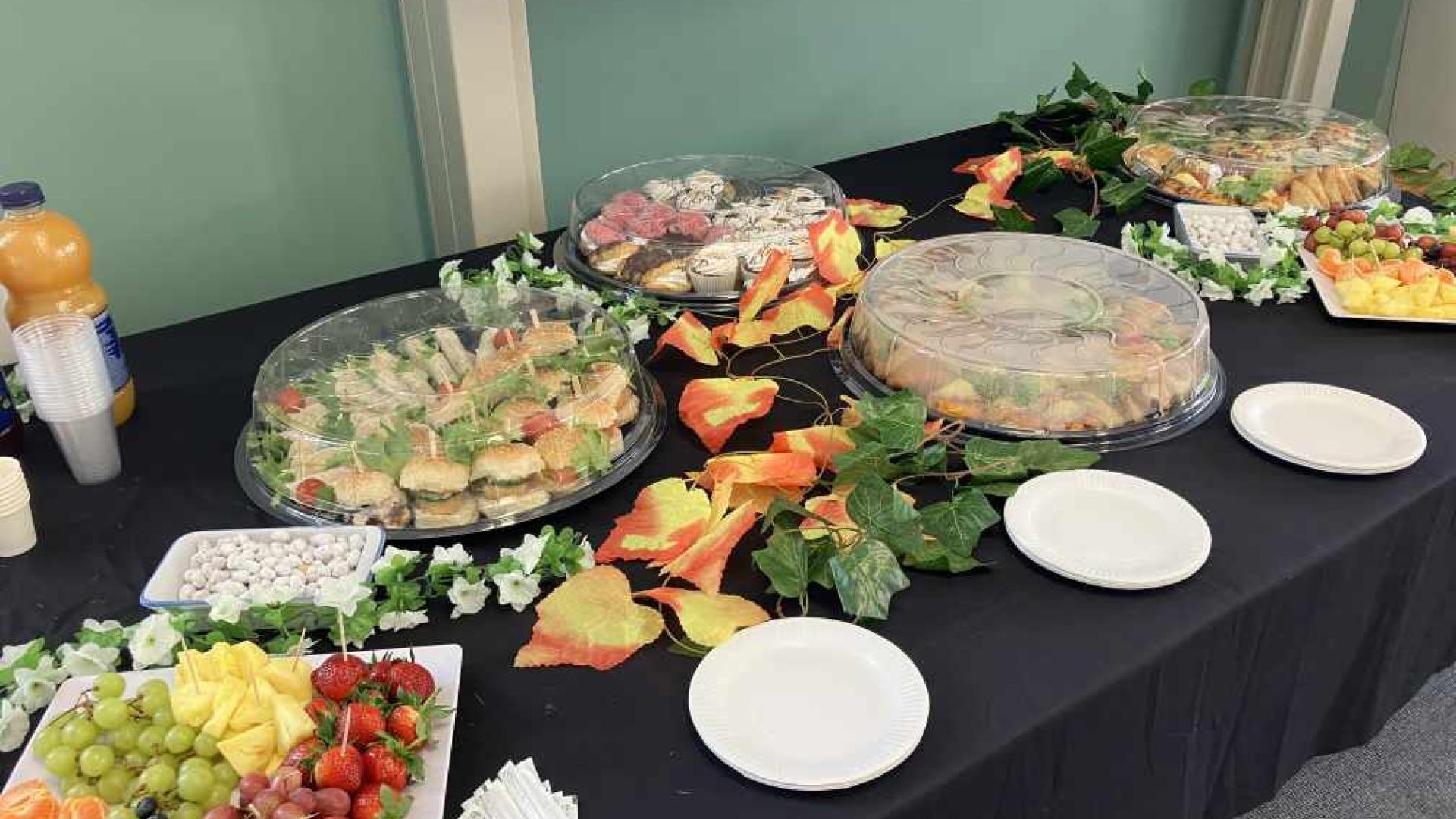 A buffet table set up for an event with platters of sandwiches, wraps, fruits, and vegetables. Decorative foliage with autumn leaves accents the presentation. A chalkboard and a bottle of orange juice are in the background.