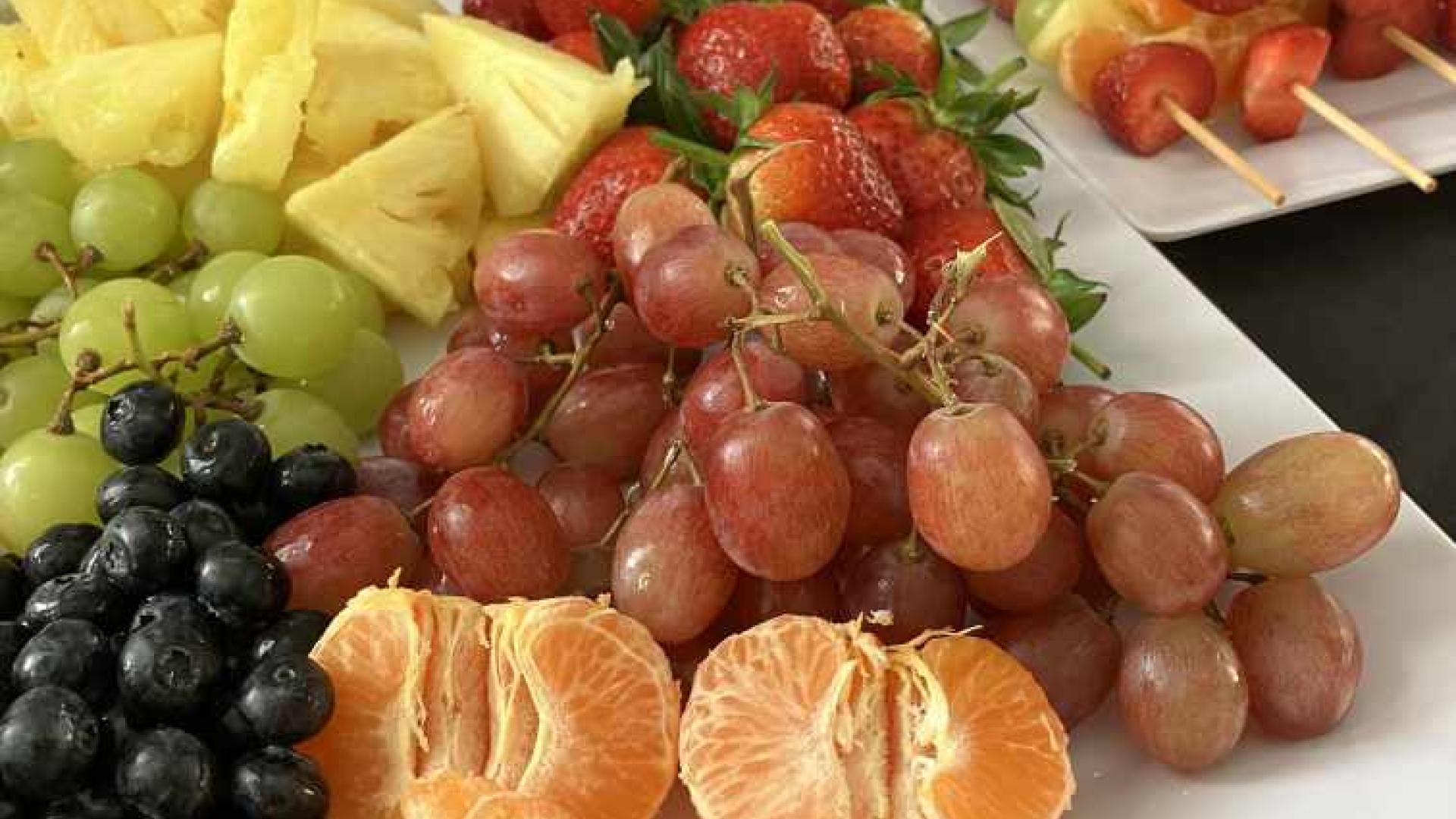 A white platter filled with a variety of fresh fruits, including red and green grapes, pineapple slices, strawberries, blackberries, and mandarin orange segments. In the background, there are fruit skewers and a bowl of nuts or small snacks on a dark surface with white flowers in the upper left corner.