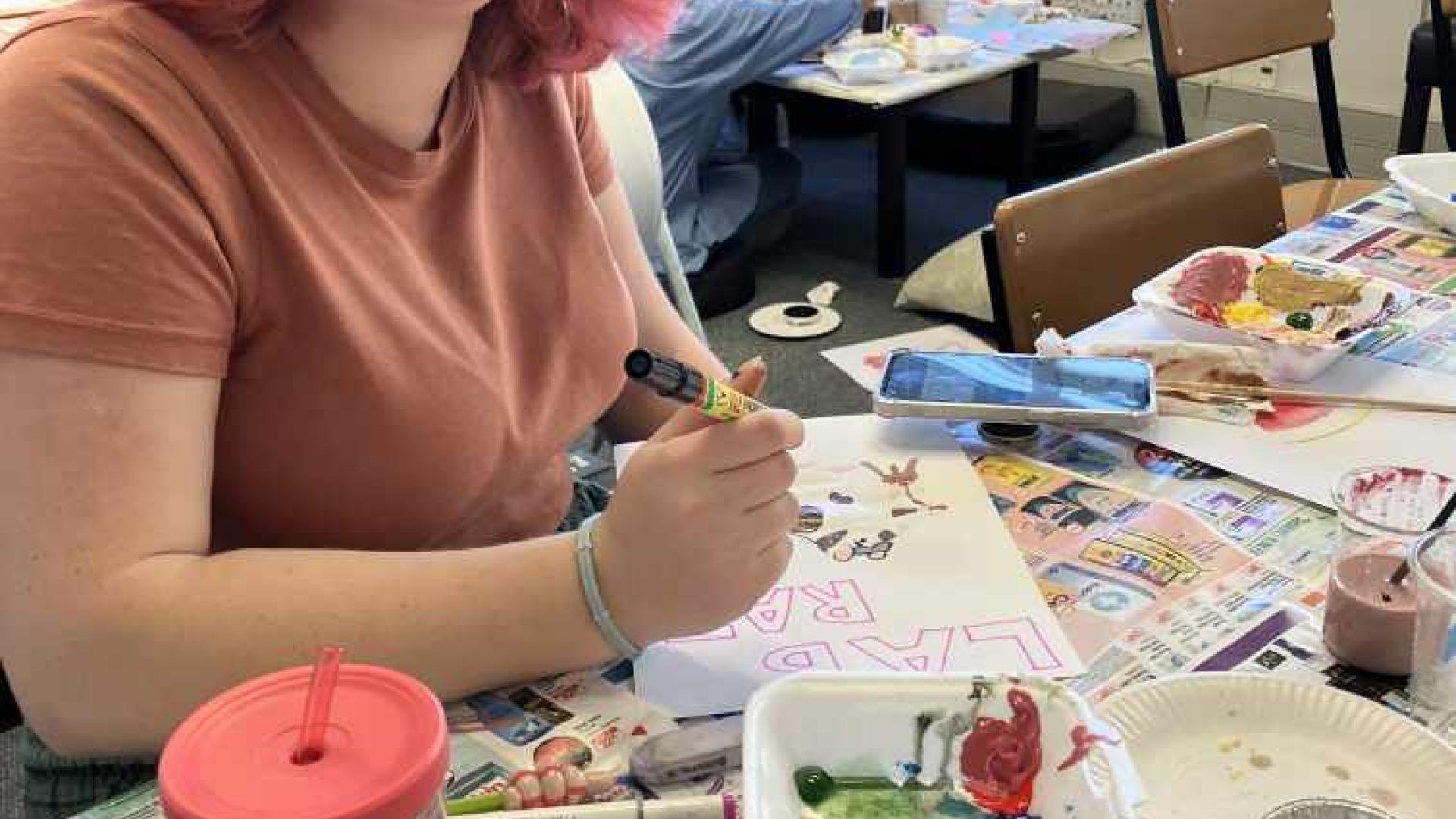 An individual is sitting at a table covered with newspapers, engaged in painting. Various art supplies, including paint palettes and brushes, are scattered around. In front of the individual is a colorful artwork featuring blue and yellow shapes on paper, suggesting an art class or workshop setting.