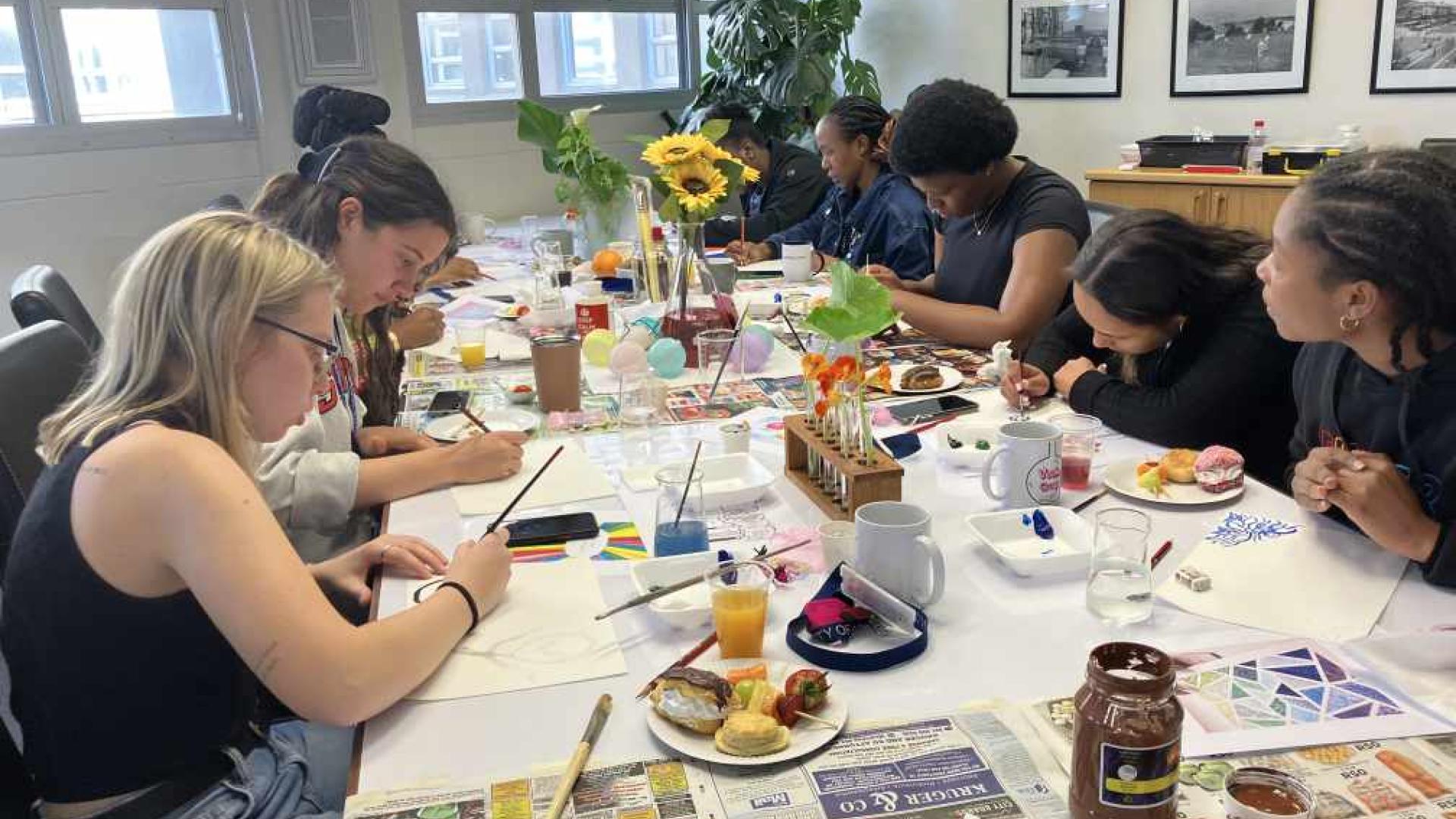 A group of individuals engaged in an art activity around a table covered with newspapers. The table has a vase of sunflowers, various painting supplies, personal items, beverages, and snacks, indicating a casual and social atmosphere.