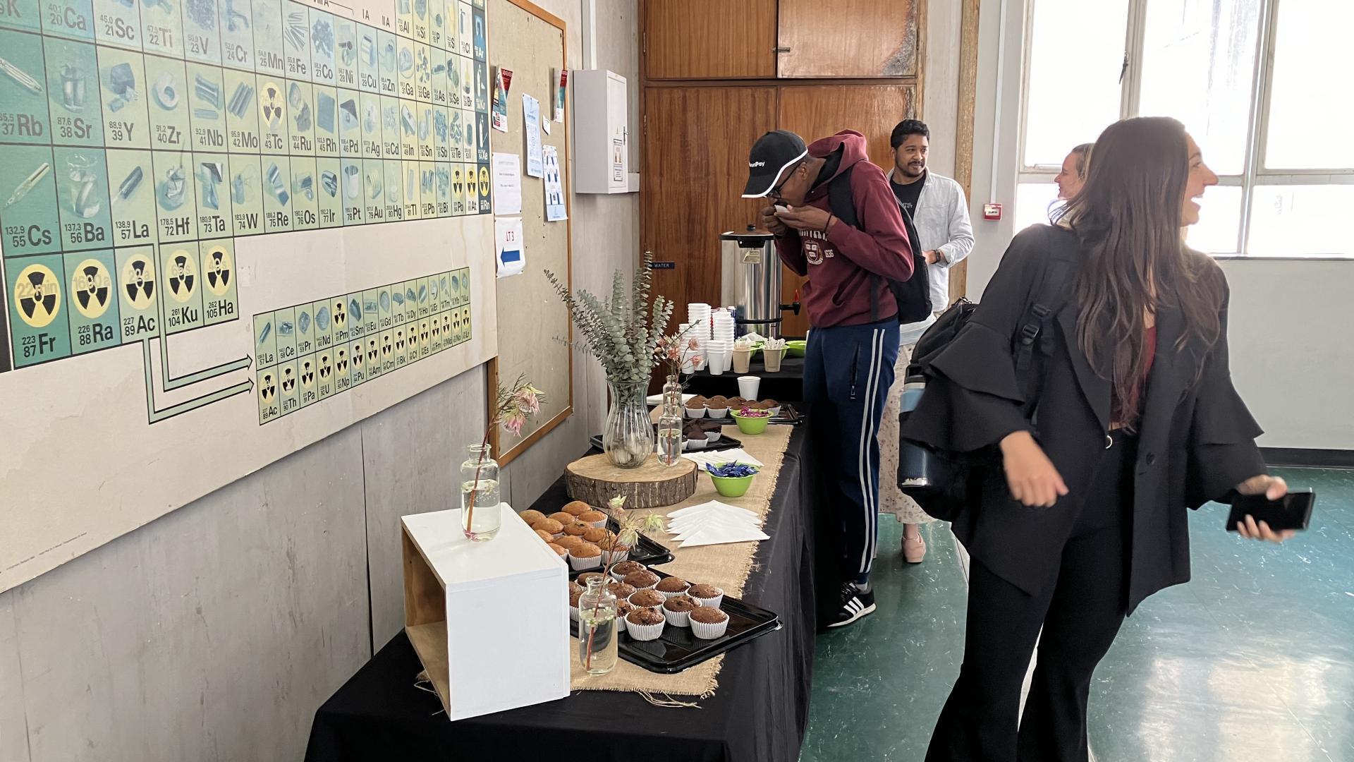 Classroom with students viewing a periodic table and a table with baked goods, likely during a science event.