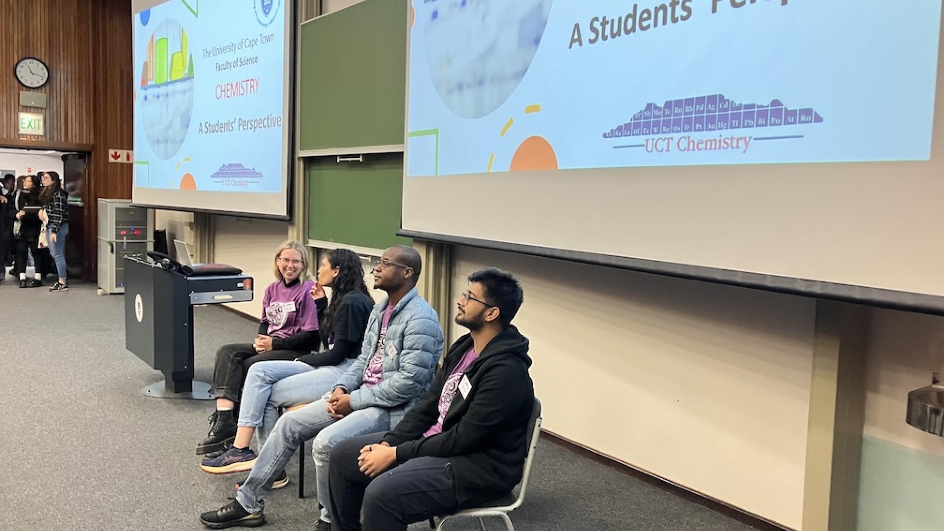 Four chemistry department postgraduate students sit on chairs in front of lecture theatre boards