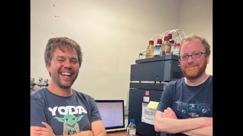 Picture of two smiling men standing in front of a mass spectrometer.
