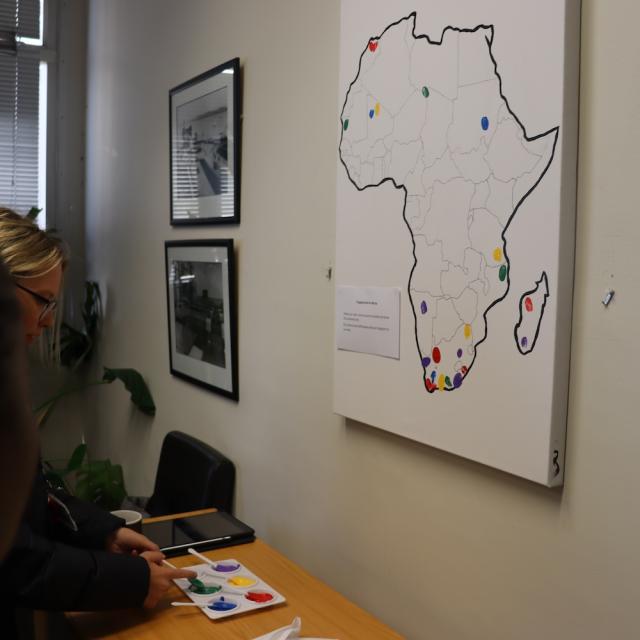 An outline of Afrika on a white canvas that is hung up on the wall with six colours paint on the table for students to show where in Afrika they come from.