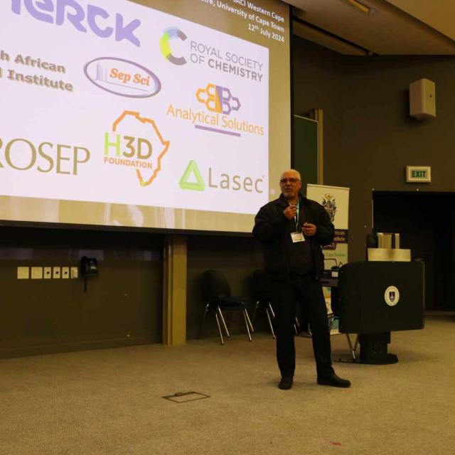 A person stands on a stage presenting to an audience. The large screen behind them displays logos of sponsors and collaborators.