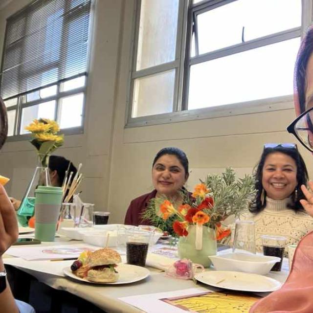 A group of people sitting around a table with food, drinks, and a vase of yellow flowers. The setting is an indoor room with windows and blinds in the background.