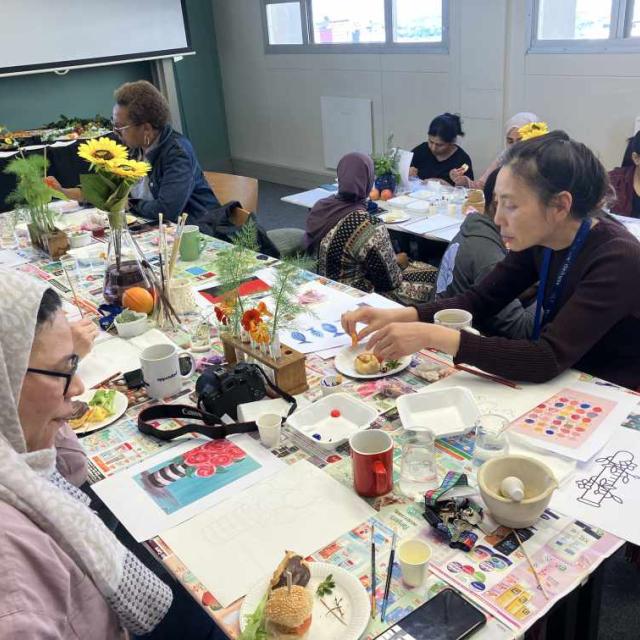 A group of individuals engaged in an art class. The tables are covered with various art supplies, including paint palettes, brushes, and watercolor papers. Some participants are painting, while others are handling different materials or having a snack. The room is bright with natural light coming in from the windows.