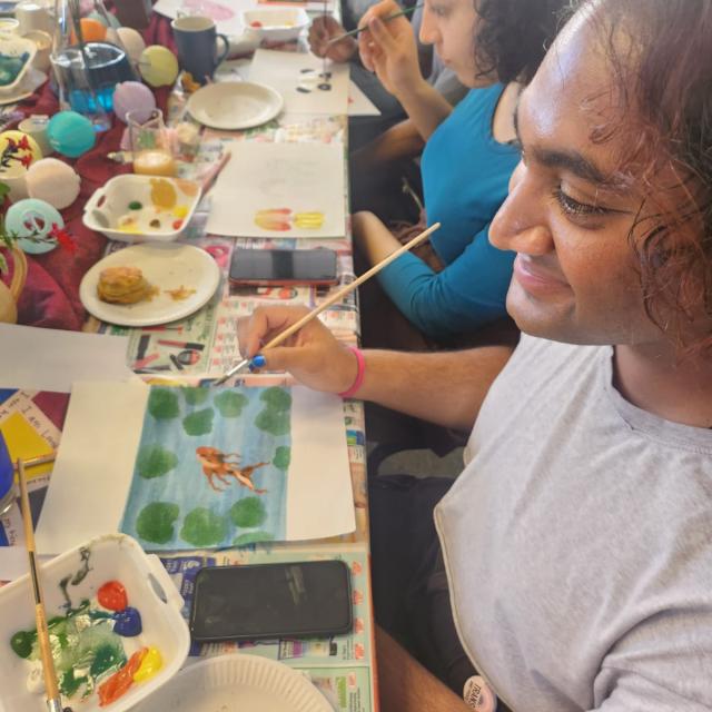 An artist’s workspace with various art supplies, including paint blobs on sheets of paper, a palette with different colors, brushes, a cup of water, and a wooden stool.