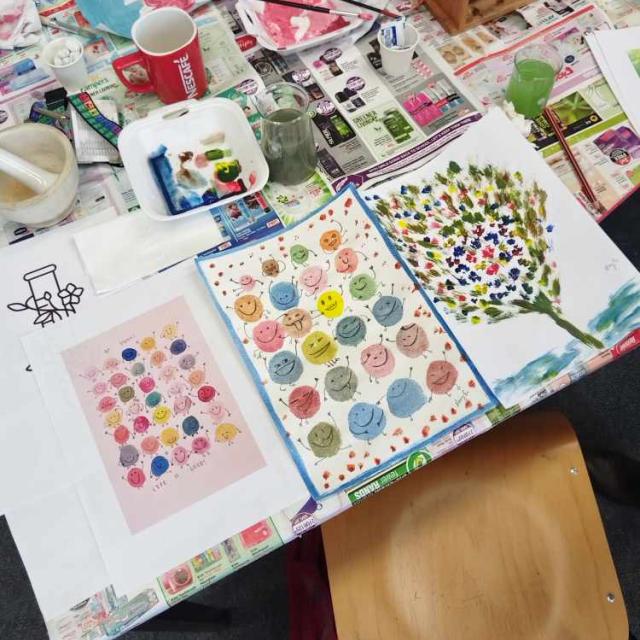 A group of people engaged in a painting activity around a table, with various art supplies like paintbrushes, paints, and palettes visible.
