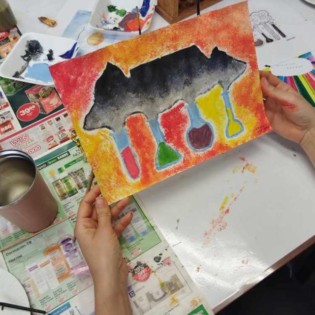 A pair of hands holding a colorful painting with an abstract design featuring a black silhouette resembling an animal, possibly a cow, with vibrant patterns and drips below. The background includes a table covered with newspapers and art supplies, suggesting an art class or creative workshop.