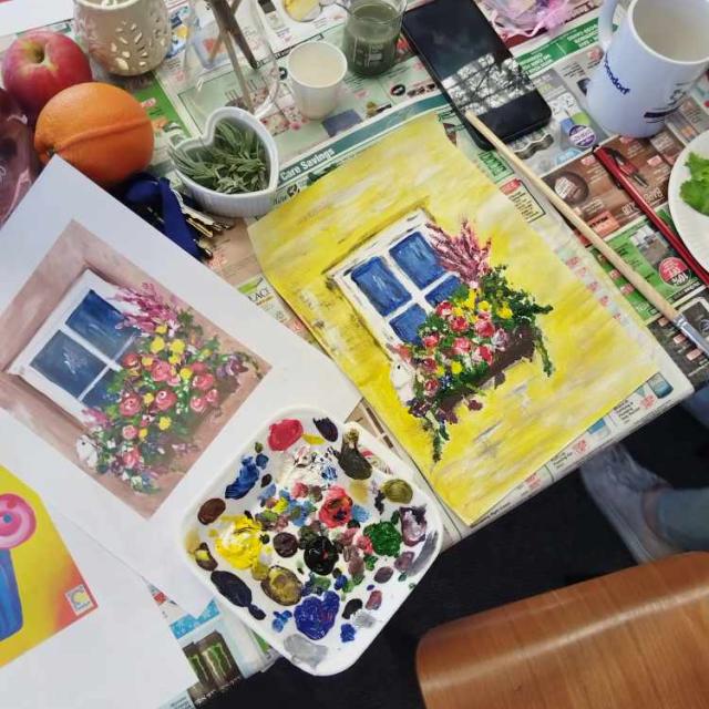A cluttered table with a watercolor painting of a window with flowers, an open book, paintbrushes, a palette with multiple colors of paint, cups, a smartphone, fruit, and other miscellaneous items, suggesting a creative workspace.