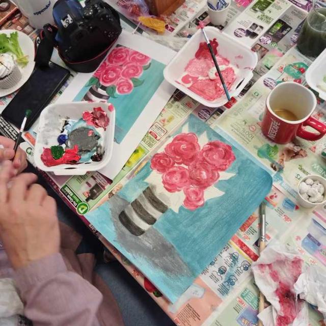 A painting in progress on a canvas, depicting pink roses in a striped vase, surrounded by painting supplies including brushes, a palette with paint, tissues, and two mugs, suggesting an art workshop or personal creative space.